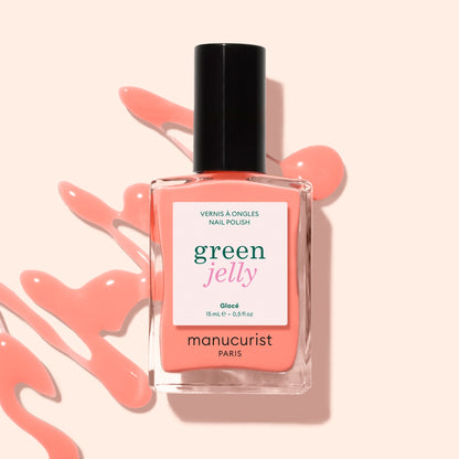 A bottle of Manucurist Glacé Jelly nail polish in a peachy-pink shade rests on a light surface, with matching ultra-glossy polish artfully spilled around it.