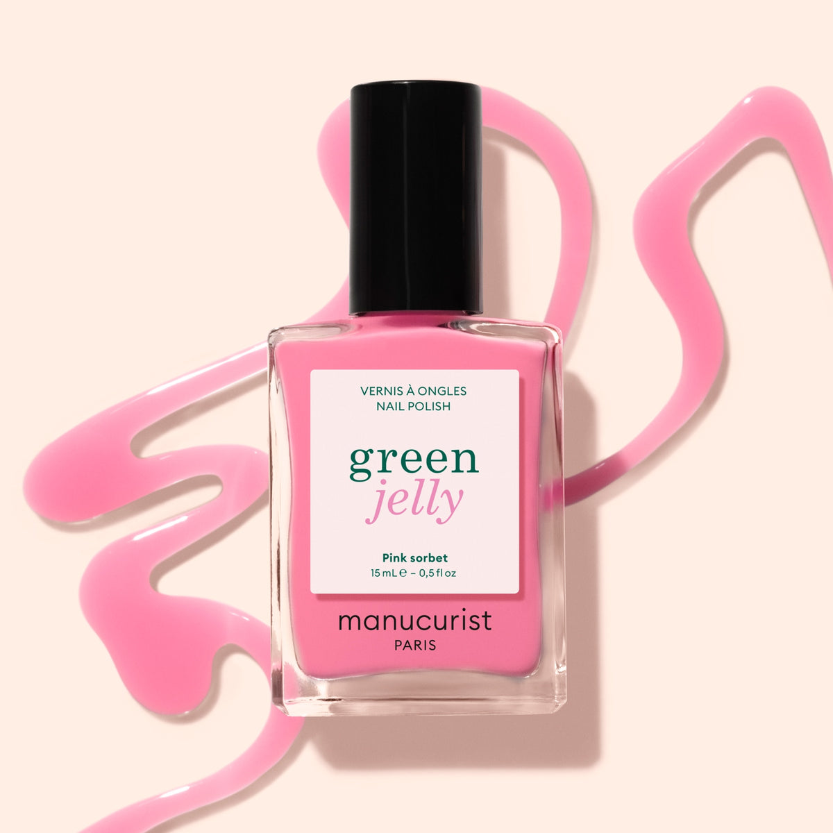 A bottle of pink nail polish labeled green jelly by Manucurist Paris sits on a light surface with pink nail polish swirled around it.