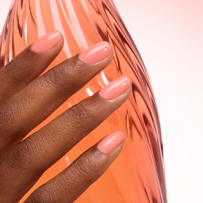 A hand with neatly manicured nails painted in Manucurist’s Glacé Jelly rests on a pink-tinted curved glass surface, with a softly lit background in matching tones for a cohesive, elegant look.
