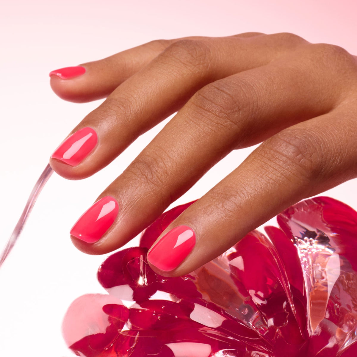 A hand with neatly manicured, glossy pink nails rests on a shiny pink glass object against a soft pink background.
