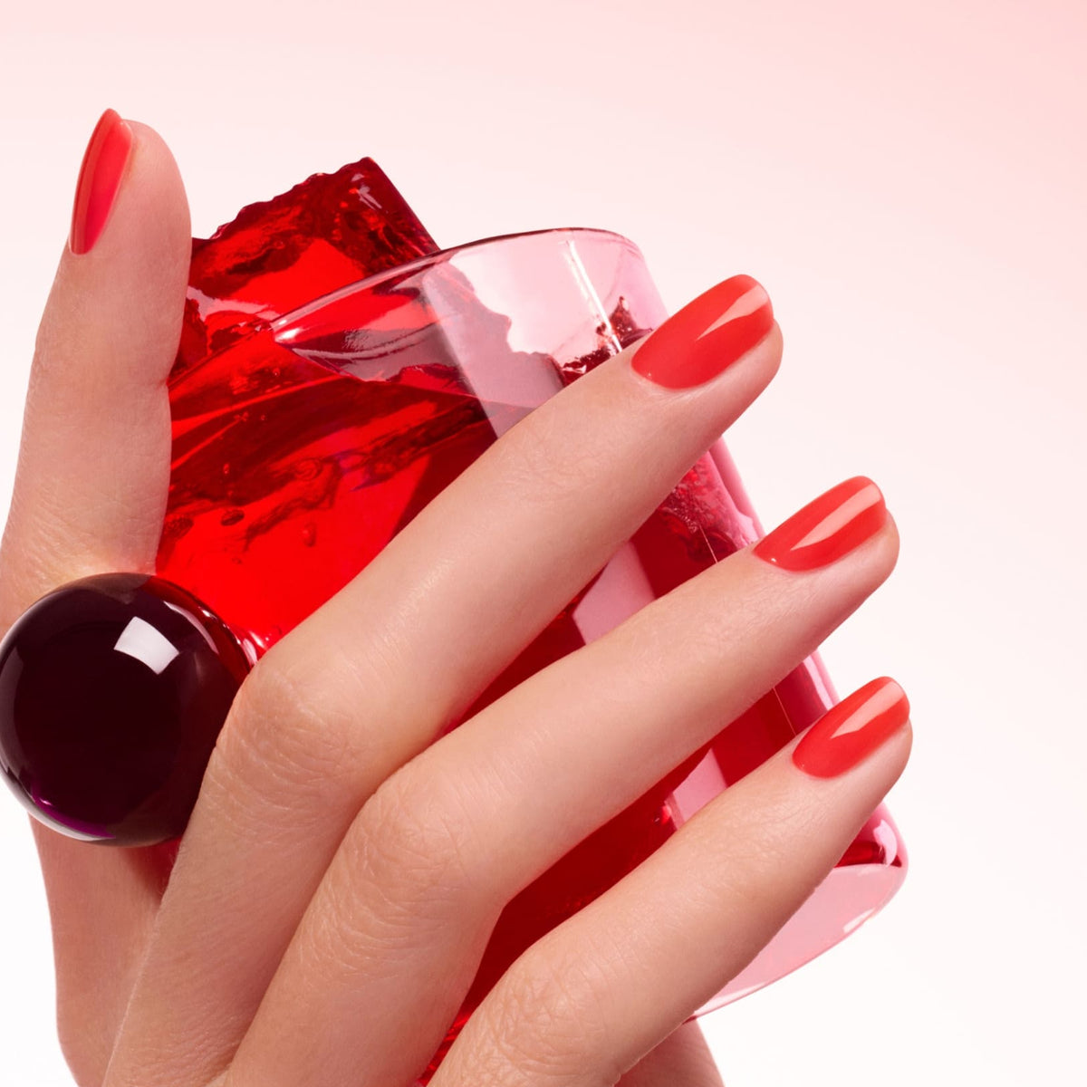 A hand with ultra-glossy, translucent red Manucurist Twizzle Jelly polish holds a transparent pink cup of red gelatin, wearing a large dark round ring against a soft pale pink gradient background.