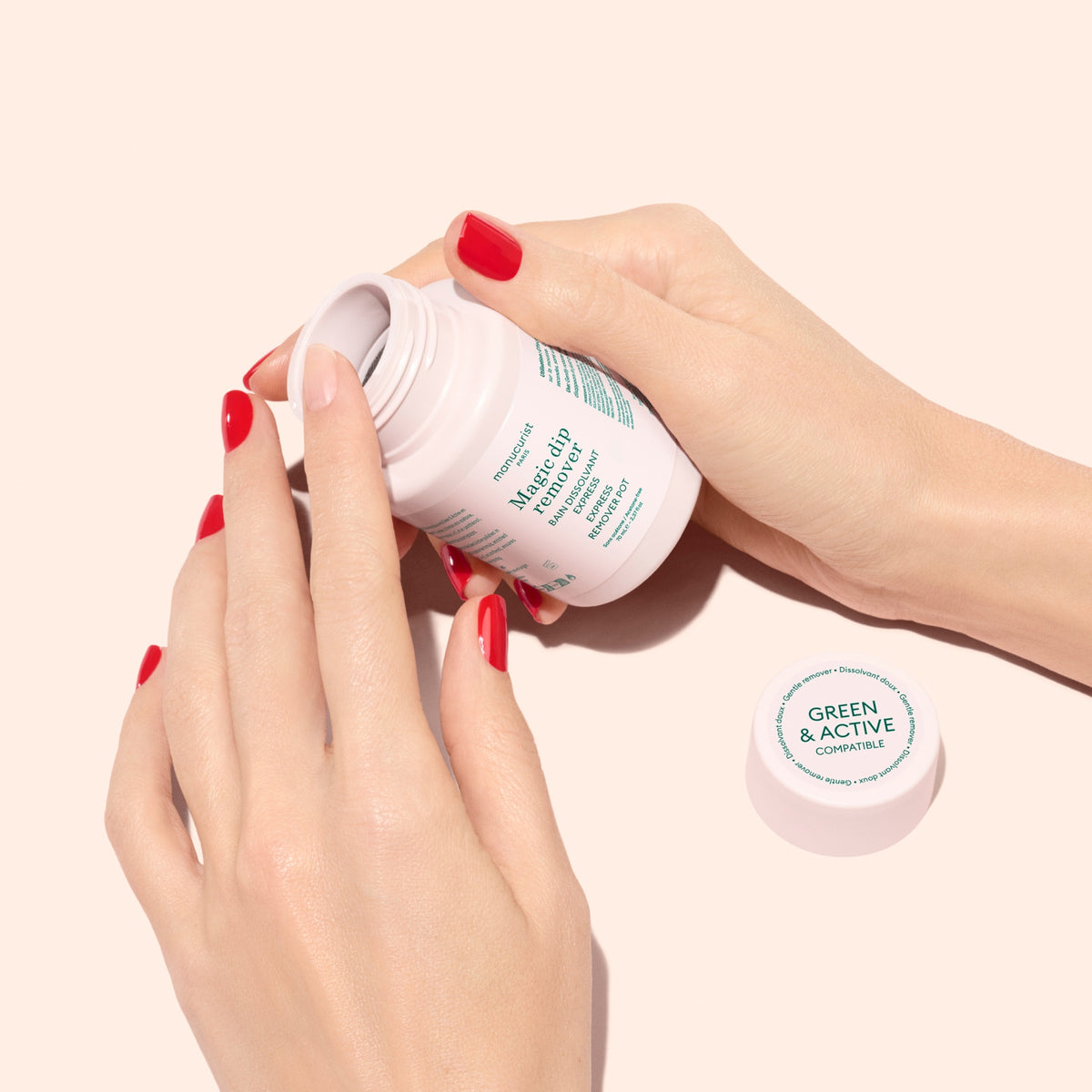 Two hands with red nail polish hold an open bottle labeled GREEN  ACTIVE and MAGNESIUM. The white bottle is tilted, and its cap lies nearby on a pale pink background.