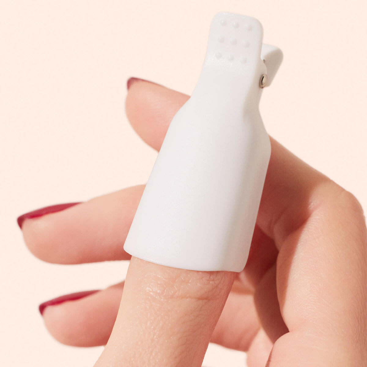 A hand with red nail polish wears a white plastic nail clip on the index finger, commonly used for removing gel nail polish.
