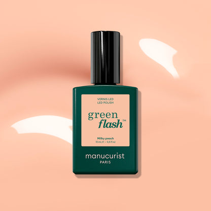 Un flacon de vernis LED Manucurist Milky Peach Green Flash™, une teinte gel pêche laiteuse aux tons beige orangé doux, présenté sur un fond pêche ponctué de reflets de lumière blanche.
