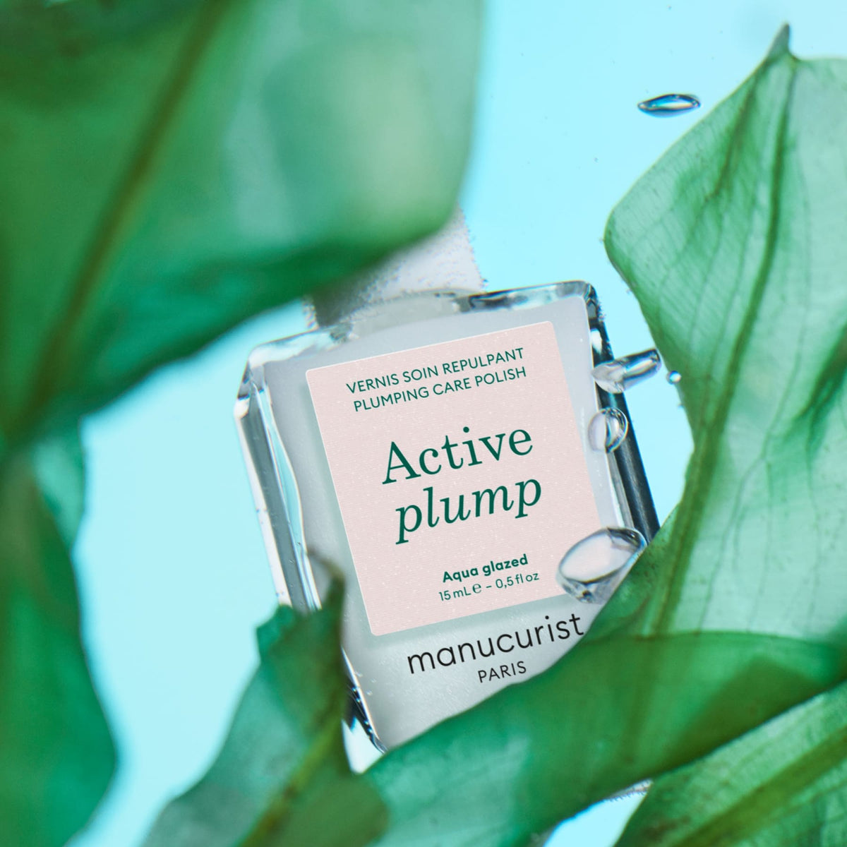 A bottle of Manucurist Active™ Plump Aqua Glazed is displayed amidst green leaves on a blue background, with its label clearly visible to emphasize the product’s promise of volumizing nail care and beautifully hydrated nails.