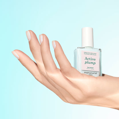 A hand with neatly manicured, hydrated nails holds a clear bottle of Manucurist Active™ Plump Aqua Glazed nail care product against a light blue background.
