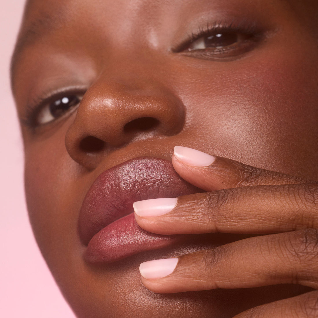A close-up of a person with dark skin touching their lips with manicured fingers. Their short nails are polished with Manucurist Active™ Blur, a light pink blurring care polish that creates a flawless matte finish. The background is softly blurred in a pink shade.