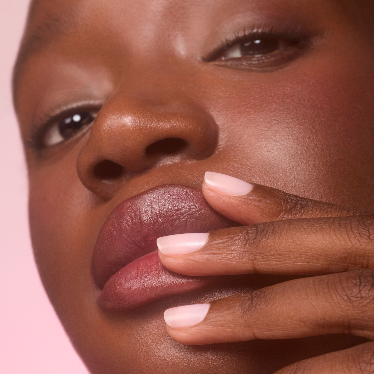 Gros plan sur une personne à la peau foncée touchant ses lèvres avec des doigts manucurés. Ses ongles courts sont vernis avec Manucurist Active™ Blur, un soin-vernis flouteur rose clair qui crée un fini mat impeccable. L’arrière-plan est délicatement flouté dans une teinte rose.