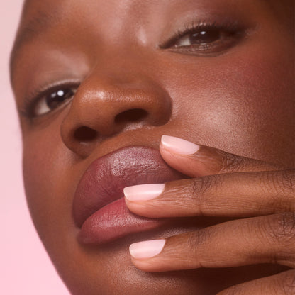 Gros plan sur une personne à la peau foncée touchant ses lèvres avec des doigts manucurés. Ses ongles courts sont vernis avec Manucurist Active™ Blur, un soin-vernis flouteur rose clair qui crée un fini mat impeccable. L’arrière-plan est délicatement flouté dans une teinte rose.