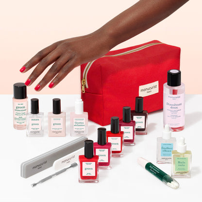 A hand with red nails reaches toward the Manucurist Super Ultimate Kit, featuring gel-effect nail polish, nail files, remover, nourishing oils, a vitaminized base coat, and a red zip pouch—all elegantly arranged on a pale surface.
