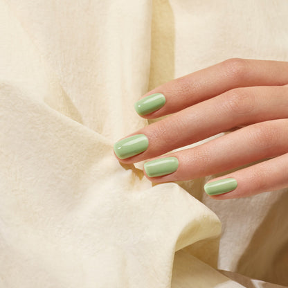 A hand with neatly manicured nails painted in Manucurist’s Milky Matcha gel polish rests gently on a cream-colored, textured fabric background, evoking creamy nuances and subtle botanical notes.