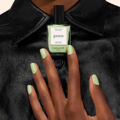 Une main ornée du vernis à ongles Milky Matcha de Manucurist tient un flacon Milky Matcha assorti sur fond de chemise en cuir noir.