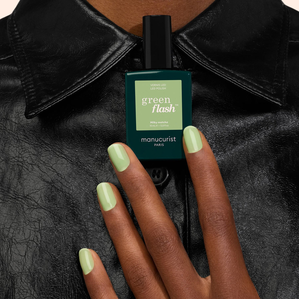 A hand with Milky Matcha gel polish—a creamy matcha green shade from Manucurist—holds a matching bottle of Manucurist polish against a black leather jacket background.