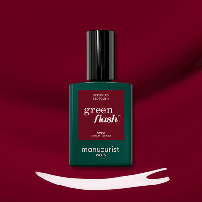 A bottle of Amour LED nail polish by Manucurist rests on a deep red surface with a brush stroke of white polish below it, highlighting the luminous burgundy hue of this signature gel polish.