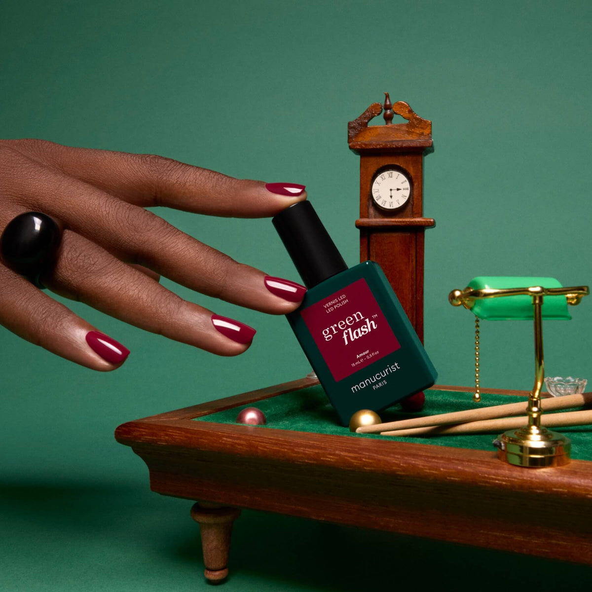 A hand with luminous burgundy nails holds a green bottle of Amour nail polish from Manucurist near a miniature pool table, with a small clock and pool cue visible on a green background.