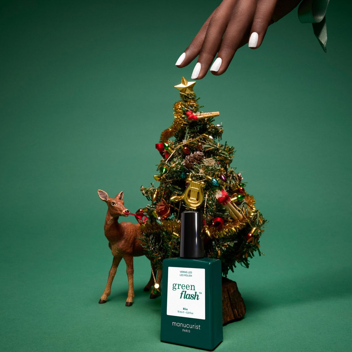 Une main aux ongles manucurés se tend au-dessus d’un sapin de Noël miniature décoré, à côté d’une petite figurine de cerf. Au premier plan, un flacon de vernis Green Flash de Manucurist en teinte Bliss se détache sur un fond vert ; ses reflets blancs irisés apportent une touche festive élégante à la scène.
