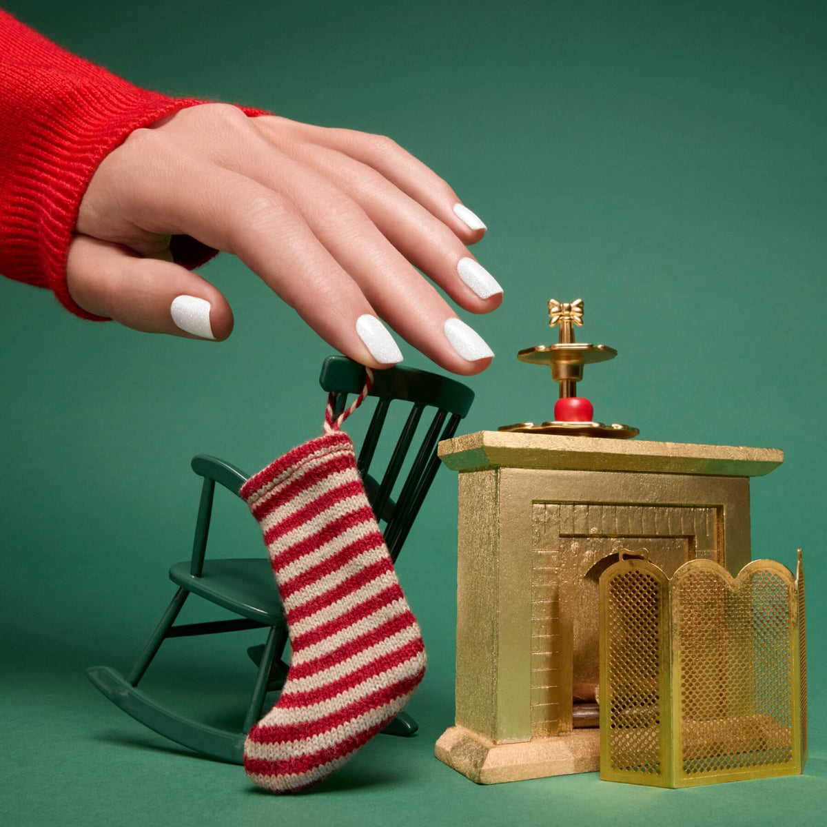 Une main aux ongles vernis d’un blanc irisé avec la teinte Bliss de Manucurist survole une petite chaise à bascule verte, une chaussette rayée rouge et blanc et une cheminée dorée miniature, sur fond vert.