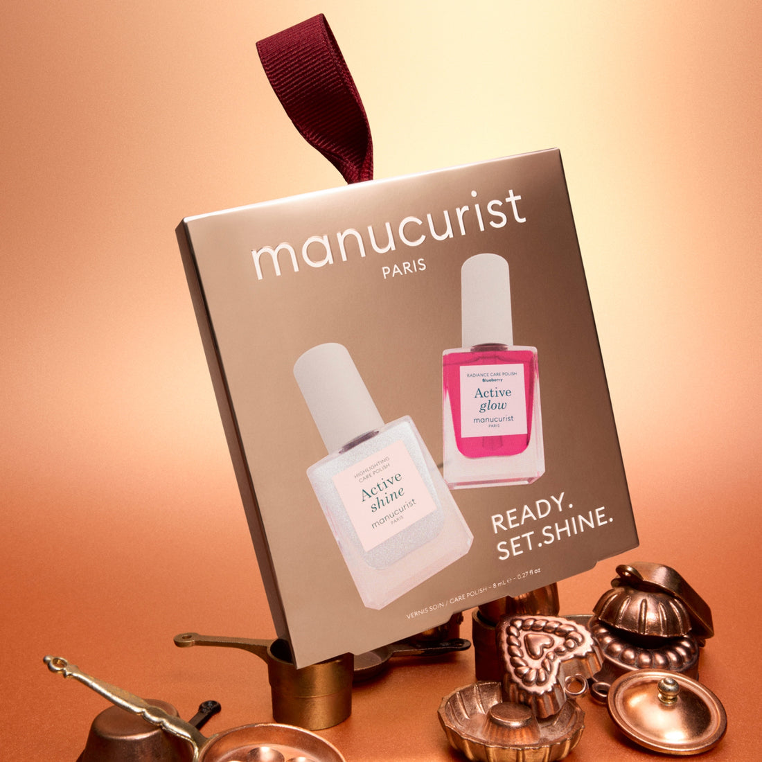 A box of Manucurist Active™ Travel-Size Duo, featuring two bottles labeled Active Shine and Active Glow, stands among small brass objects on a brown surface with a gold-to-peach gradient background—perfect for nail care enthusiasts.