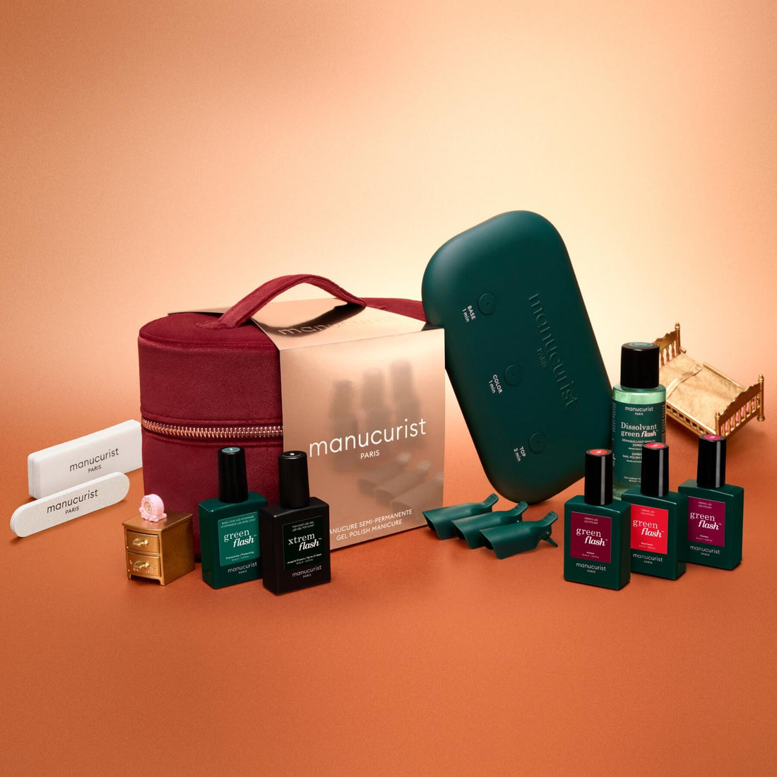 The Green Flash™ Starter Kit Gift Set by Manucurist is arranged on a peach-colored surface, featuring nail polishes, a maroon case, a green LED lamp, nail files, and accessories—perfect for achieving long-lasting salon-style manicures at home with an easy gel polish application.
