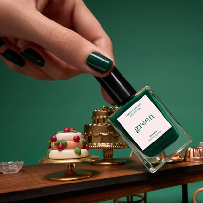Une main aux ongles vernis en vert forêt profond avec la teinte Emerald de Manucurist tient un flacon de vernis semi-permanent LED Green Flash de Manucurist au-dessus d’une table garnie de gâteaux et de desserts miniatures, le tout sur un fond vert intense.