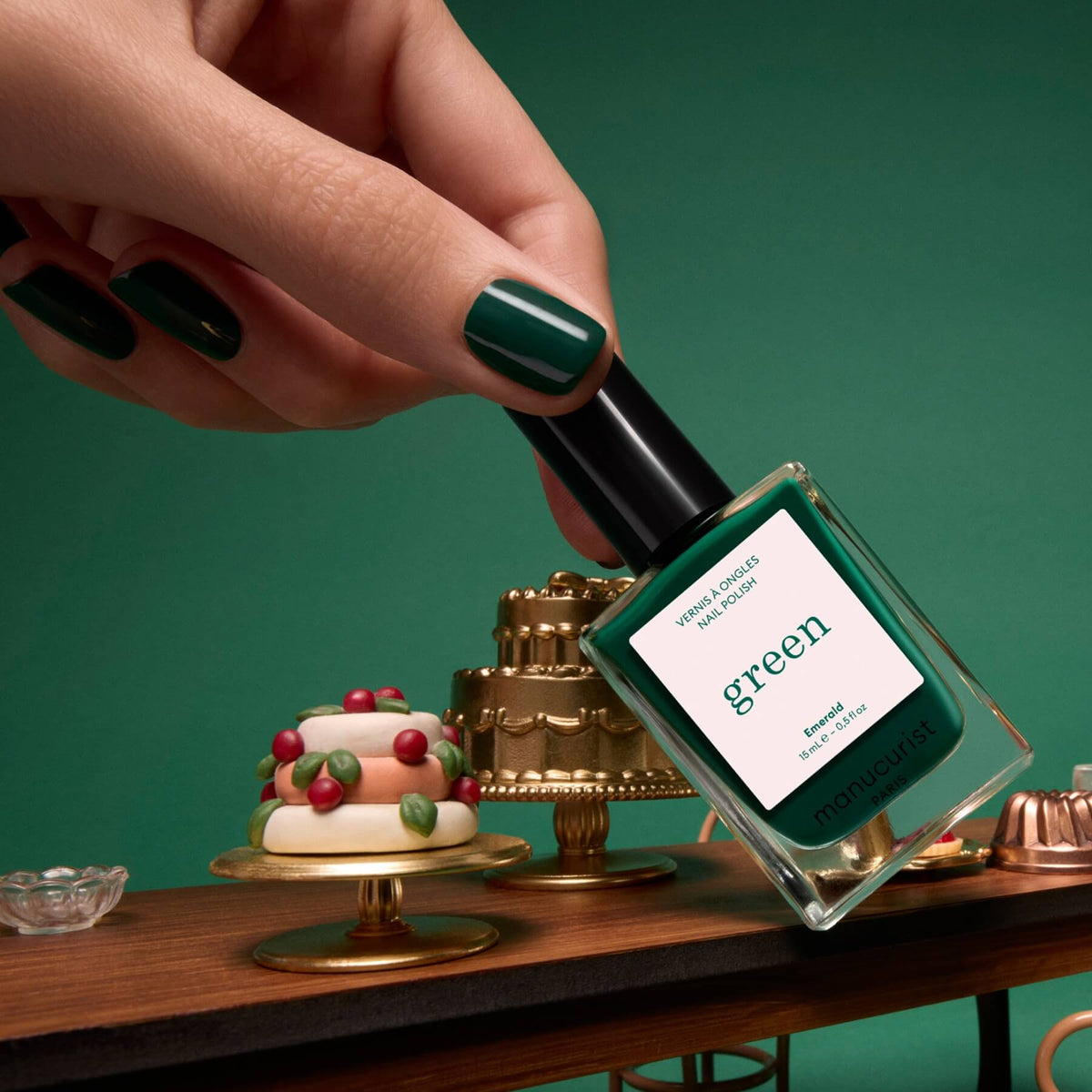 A hand with green-painted nails, featuring Manucurist’s Emerald nail polish, holds a bottle of deep forest green polish over a table with miniature cakes and desserts against a green background.