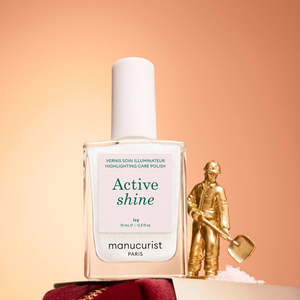 A bottle of Manucurist Active Shine nail polish stands next to a small gold figurine of a worker with a shovel, against a peach gradient background.