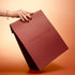 A hand with painted nails and a bracelet holds the large Holiday Gift Bag - M from Manucurist, featuring a matte burgundy finish, against a gradient peach background.