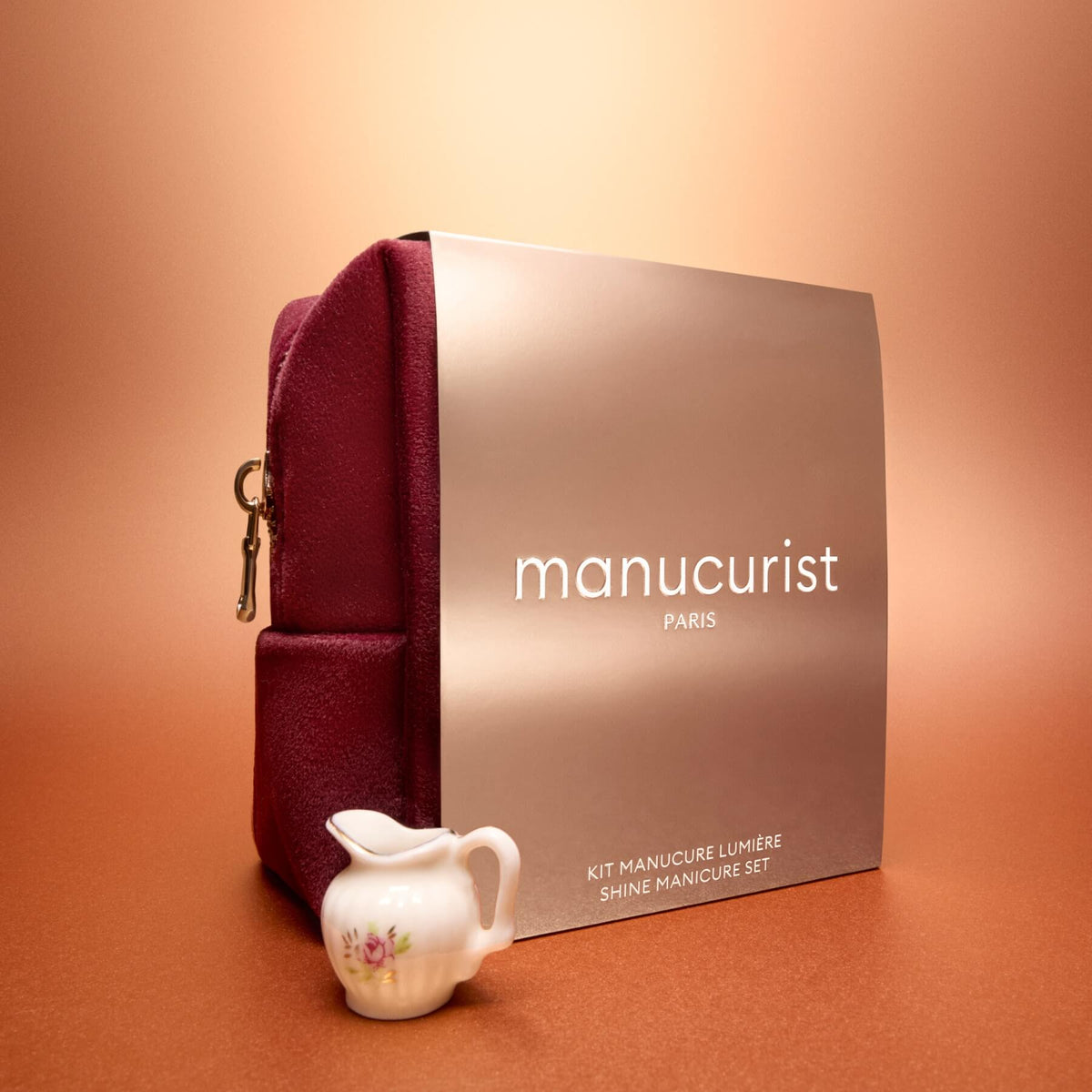 Un Active™ Gift Set Holiday de Manucurist, composé d’une pochette bordeaux et d’une boîte métallisée portant l’inscription « Manucurist Paris », est présenté sur une surface orange, accompagné à l’avant d’un petit pot à lait en porcelaine blanche décoré de fleurs.