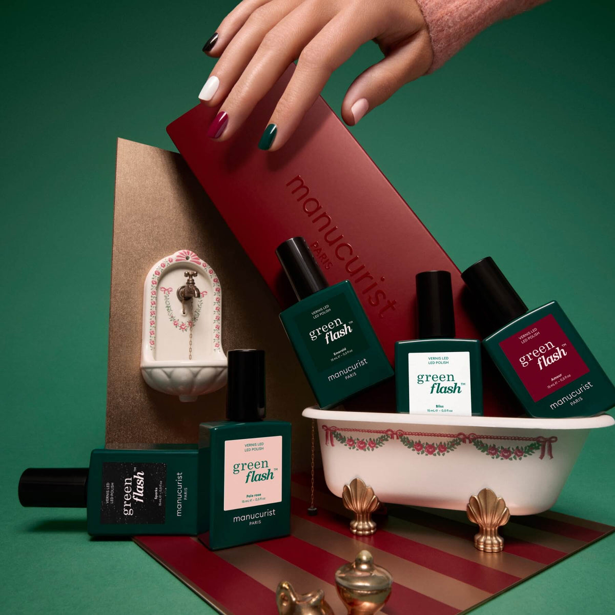Une main aux ongles multicolores survole un coffret cadeau portant le nom de Manucurist. Six flacons de vernis à ongles des Collections de couleurs sélectionnées, inspirées des palettes de couleurs Green Flash, sont disposés dans et autour d’une petite baignoire décorative, tandis qu’un minuscule lavabo orné se trouve à l’arrière-plan. Les tons verts et rouges mettent en valeur les dernières tendances manucure.