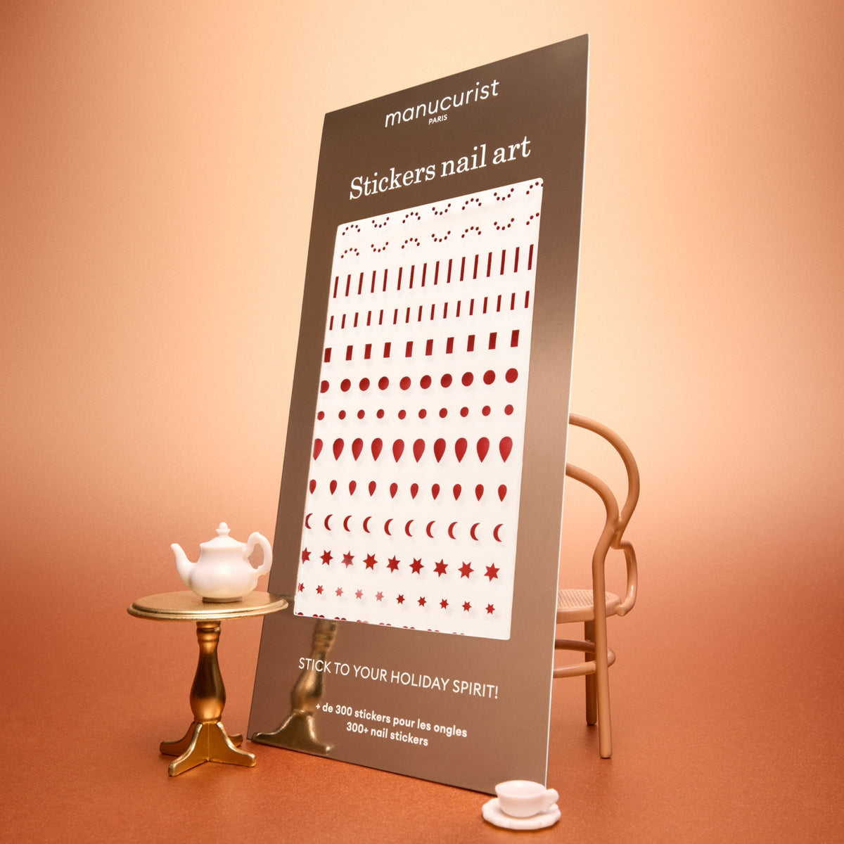Un présentoir de stickers Nail Art Holidays de Manucurist montre des motifs festifs rouges sur des planches blanches, parfaits pour créer des manucures de fête. La scène se déroule sur une surface couleur pêche, avec une petite table portant une théière et une tasse, et une chaise en arrière-plan.