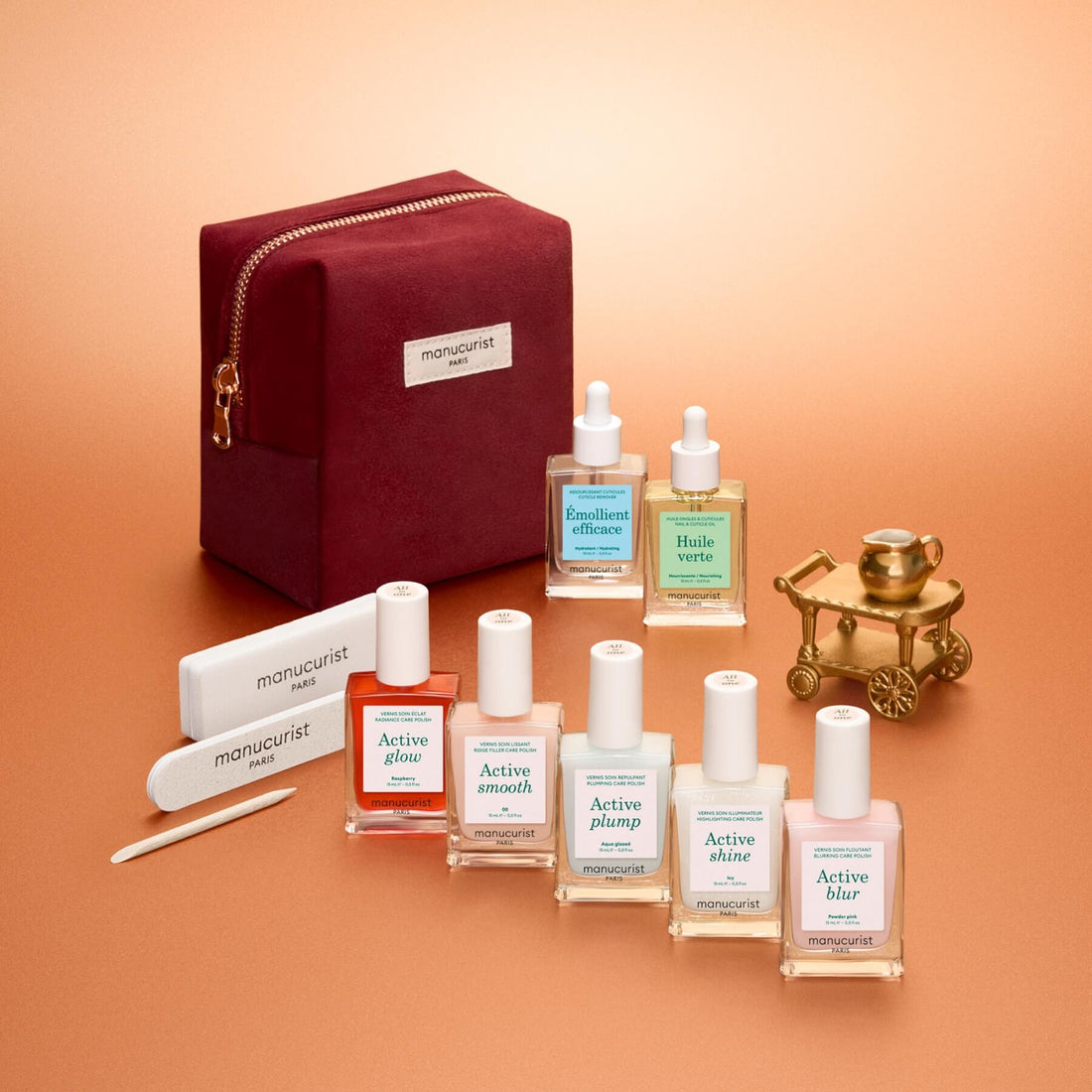 The Manucurist Active™ Super Starter Gift Set features a vibrant red pouch, six nail care bottles, a nail file, and a cuticle stick—all elegantly displayed on a peach background alongside a decorative gold carriage. Each item is labeled with effective nail treatments to help you achieve beautiful nails, making this set an ideal holiday gift for manicure enthusiasts.
