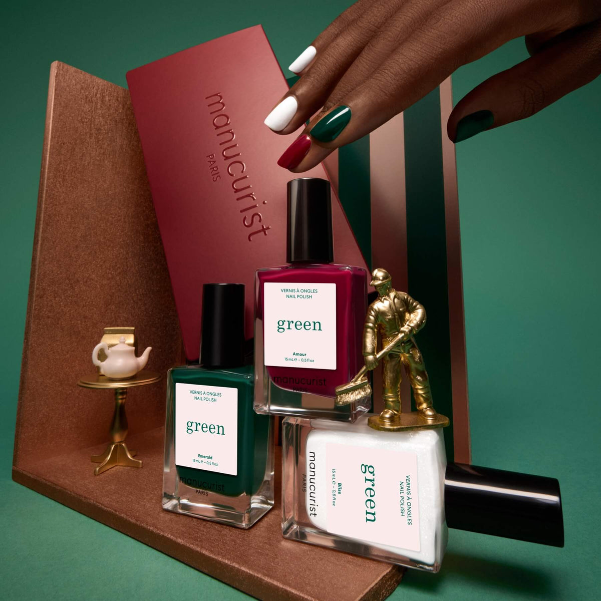 Une main aux ongles vernis tient un coffret bordeaux portant l’inscription « Trios sélectionnés » de Manucurist. À ses côtés, trois flacons de vernis Manucurist, une figurine miniature de golfeur doré et un petit service à thé sur un support doré sont disposés avec soin sur des plateformes géométriques pour mettre en valeur cette élégante collection de palettes de couleurs inspirées des tendances manucure.