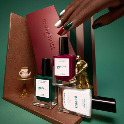 Une main aux ongles vernis tient un coffret bordeaux portant l’inscription « Trios sélectionnés » de Manucurist. À ses côtés, trois flacons de vernis Manucurist, une figurine miniature de golfeur doré et un petit service à thé sur un support doré sont disposés avec soin sur des plateformes géométriques pour mettre en valeur cette élégante collection de palettes de couleurs inspirées des tendances manucure.