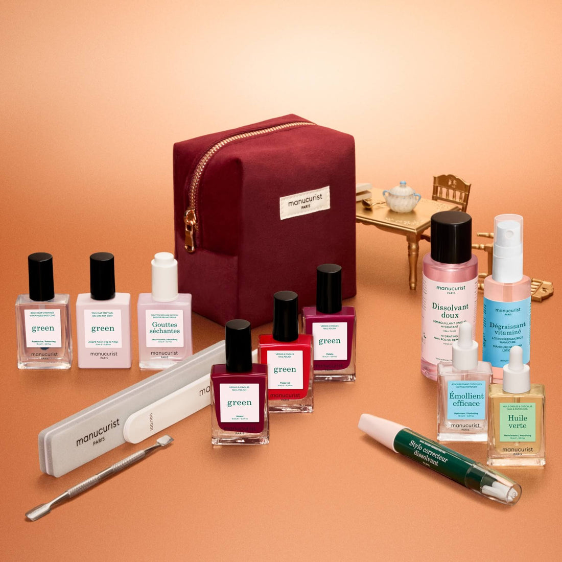 The Manucurist Green™ Ultimate Kit Gift Set is elegantly displayed, featuring a burgundy manicure bag surrounded by gel effect polish, naturally derived nail polishes, treatments, nail files, and professional manicure tools. In the background, a miniature golden table and chair with a teapot and cup add charm to the scene. All products are thoughtfully arranged—ideal for creating an at-home manicure experience.