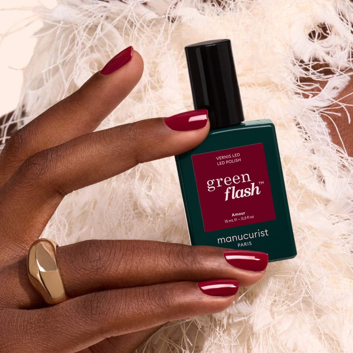A hand with luminous burgundy nail polish holds a green bottle of Amour gel polish by Manucurist, with white feathers in the background.