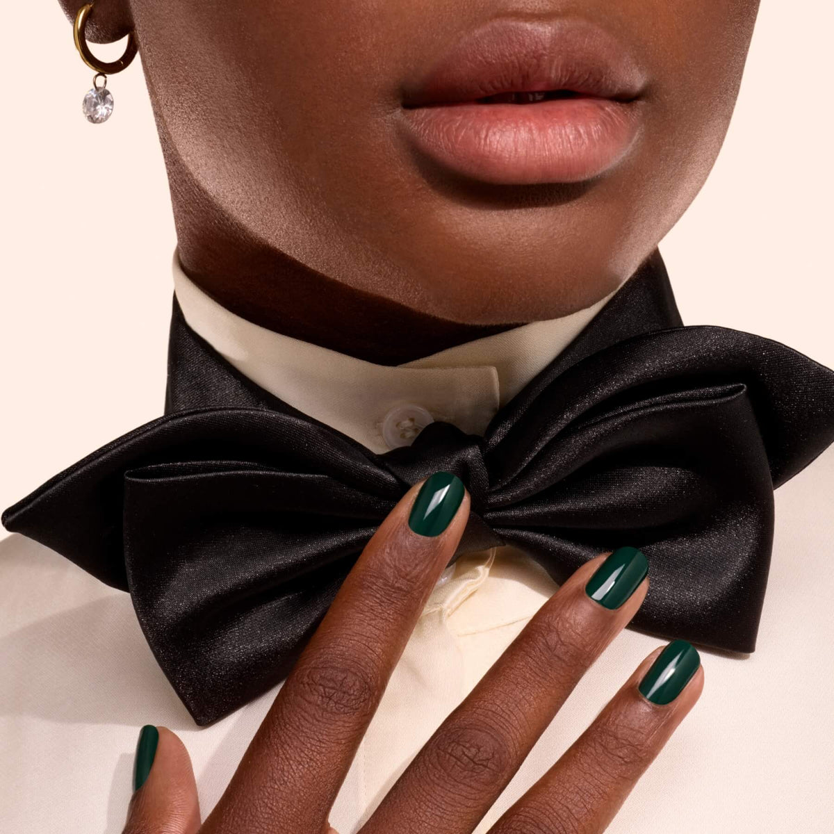 Une personne à la peau foncée porte un nœud papillon noir, une chemise crème et une boucle d’oreille dorée. Ses ongles, vernis avec la teinte Emerald de Manucurist, effleurent le nœud papillon.