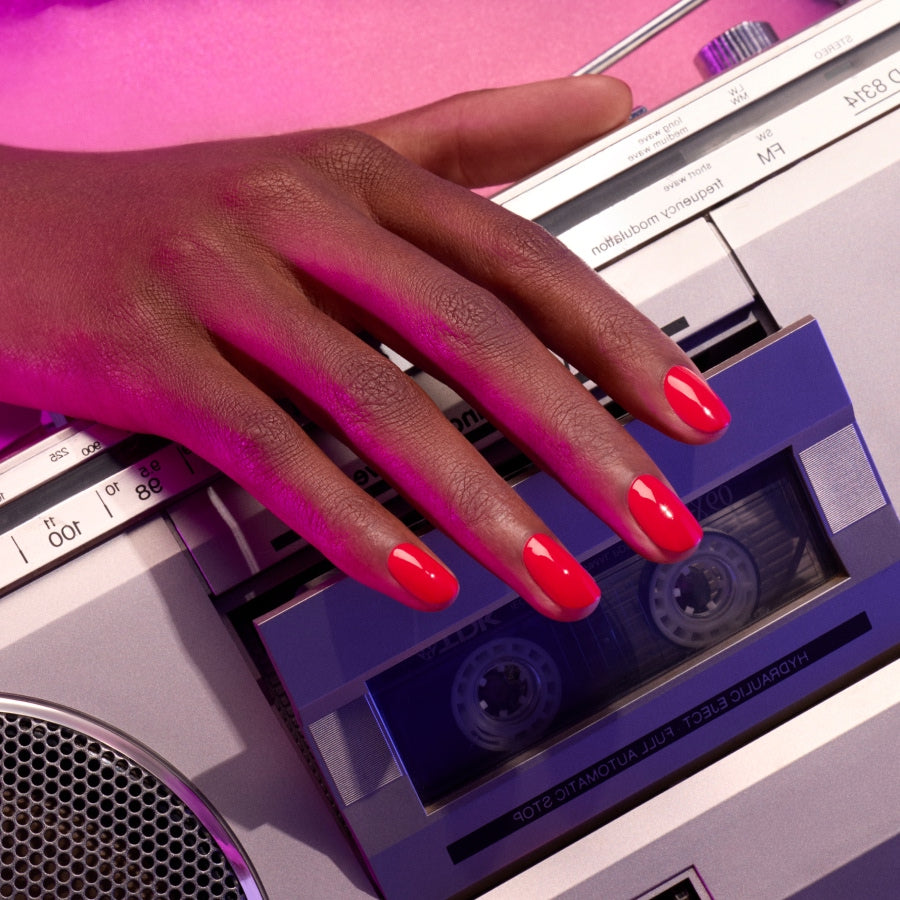 A hand with glossy, vibrant Spicy red nail polish by Manucurist rests on a silver and blue cassette player, bathed in pinkish light for an electric vibe.