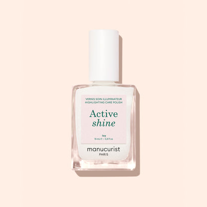 A clear glass bottle of Manucurist Active™ Shine Icy illuminating nail polish with a white cap, photographed against a pale peach background.