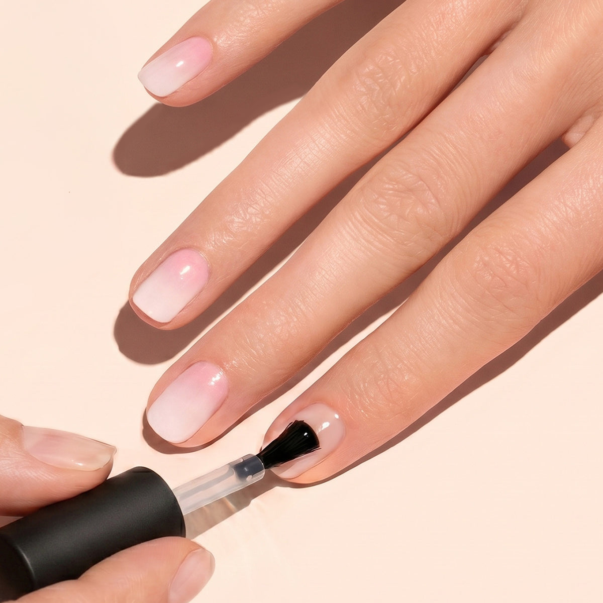 A hand is shown getting a clear nail polish applied on natural, pink-toned, manicured nails with a brush. The background is a light, neutral color.