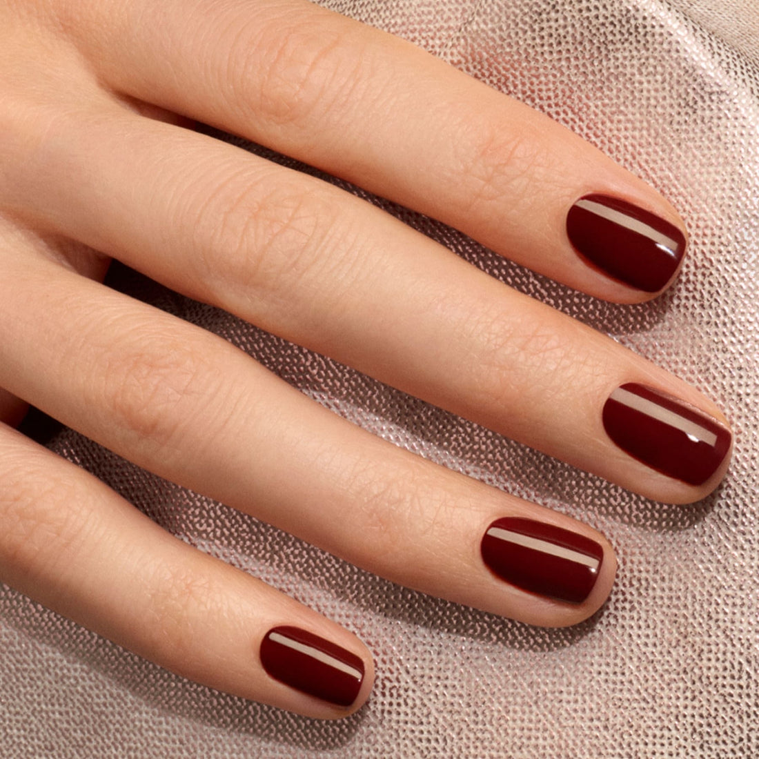 Gros plan sur une main aux ongles soignés, vernis en Grenat de Manucurist, un vernis gel rouge bordeaux brillant, posée sur un tissu texturé clair.