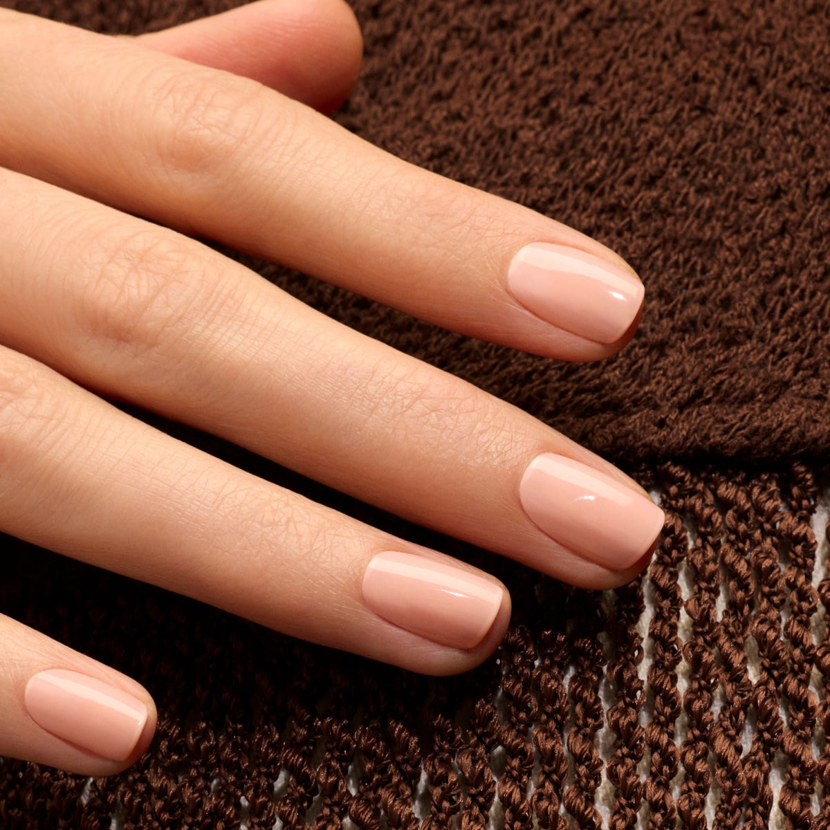 Gros plan d’une main aux ongles soigneusement manucurés, vernis avec le gel polish Milky Peach de Manucurist, posée sur un tissu marron texturé.