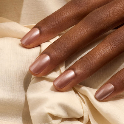 Une main à la peau brun moyen repose sur un tissu couleur crème. Les ongles sont soigneusement manucurés et vernis avec la teinte Sandstone de Manucurist, un brun latte scintillant qui leur donne une finition élégante et brillante.
