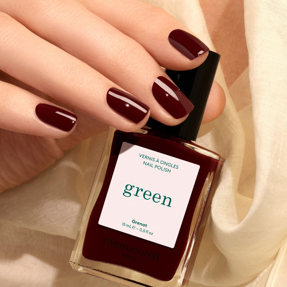 A hand with neatly manicured, maroon red nails holds a bottle of Manucurist Grenat nail polish. The background features a soft, cream-colored fabric.