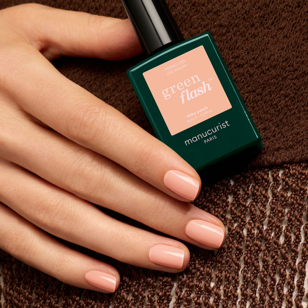 Une main aux ongles courts, brillants et rose clair — rappelant un vernis gel pêche laiteux — tient un flacon de vernis à ongles Manucurist Milky Peach, posée sur un tissu marron texturé.