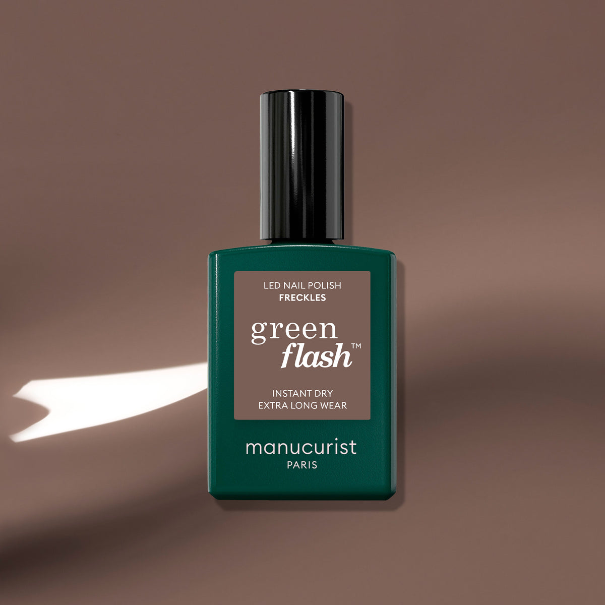 A bottle of Manucurist Freckles gel polish sits on a smooth, wood-toned brown surface with a white light shadow. The label reads: Freckles, Instant Dry, Extra Long Wear.