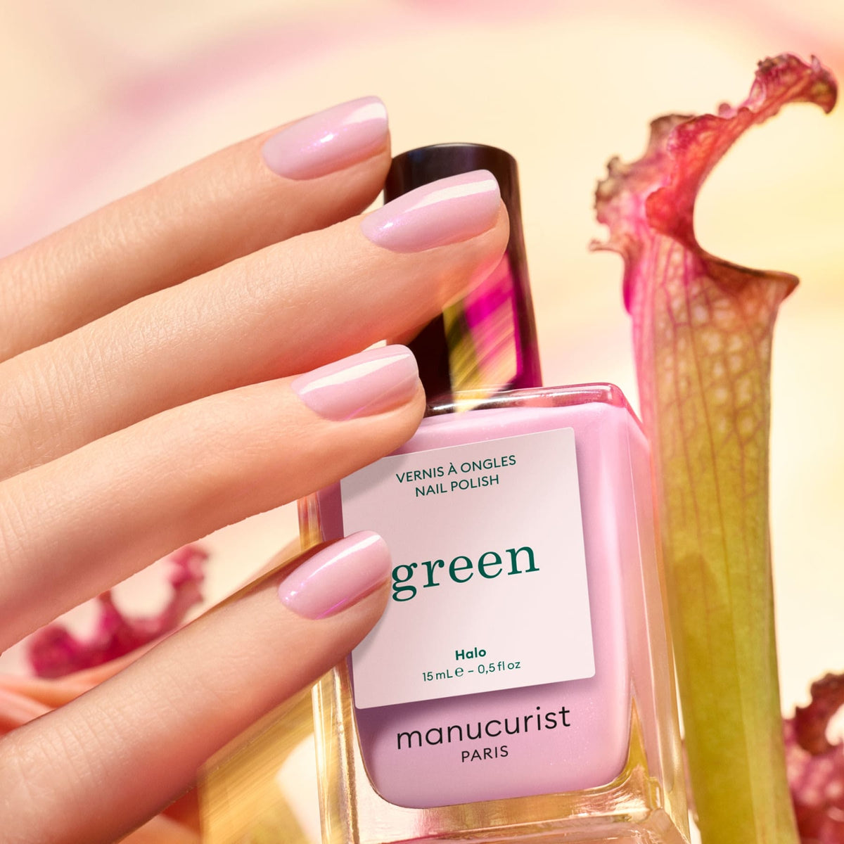 Une main aux ongles mauve rose clair laiteux et brillants tient un flacon rose "Halo" de Manucurist. Une fleur rose et une tige verte sont floues à l&
