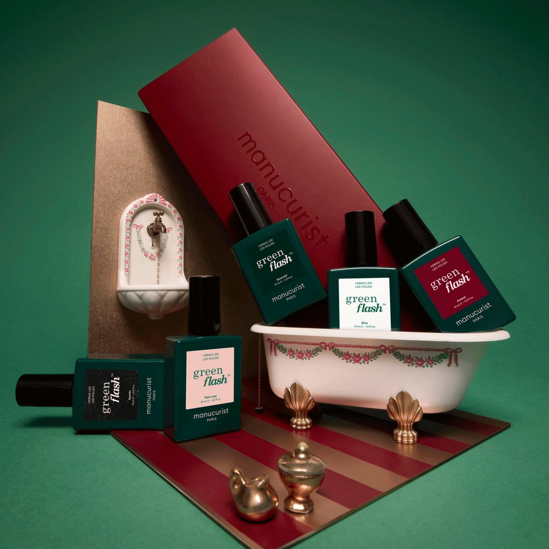 Plusieurs flacons du lot de 5 personnalisable Manucurist sont disposés autour d’une baignoire et d’un lavabo miniatures de style vintage, avec une boîte rouge Manucurist à l’arrière-plan, le tout sur un fond vert.