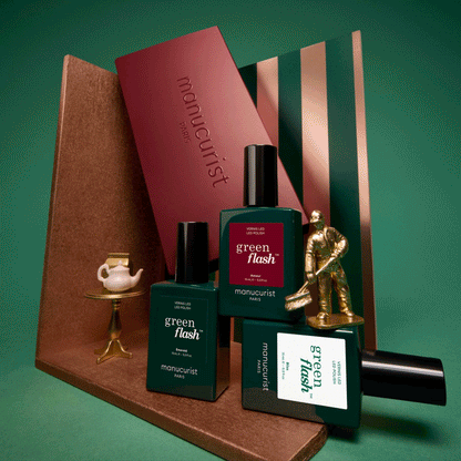 Three green bottles labeled Customizable Trio by Manucurist are displayed alongside a small gold teapot set, a gold statuette, and geometric props against a green background, creating a stylish, modern arrangement.