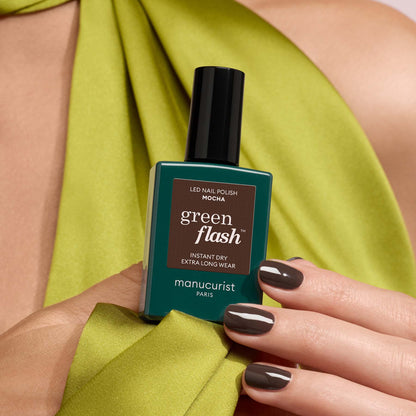 A hand with glossy dark brown Manucurist Mocha nail polish holds a bottle of the same shade, showcasing cool undertones. The person is wearing a lime green garment draped over their shoulder.