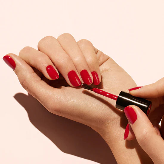 A person painting the nails on their left hand with bright red nail polish, holding the brush in their right hand. The nails are neatly manicured and the background is a soft, light color.